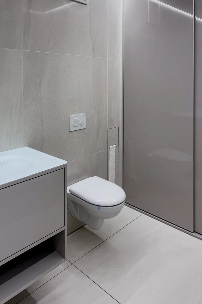 Toilette in modernem Bad mit grauen Fliesen, Waschtischunterschrank und Duschabtrennung mit Glastür.