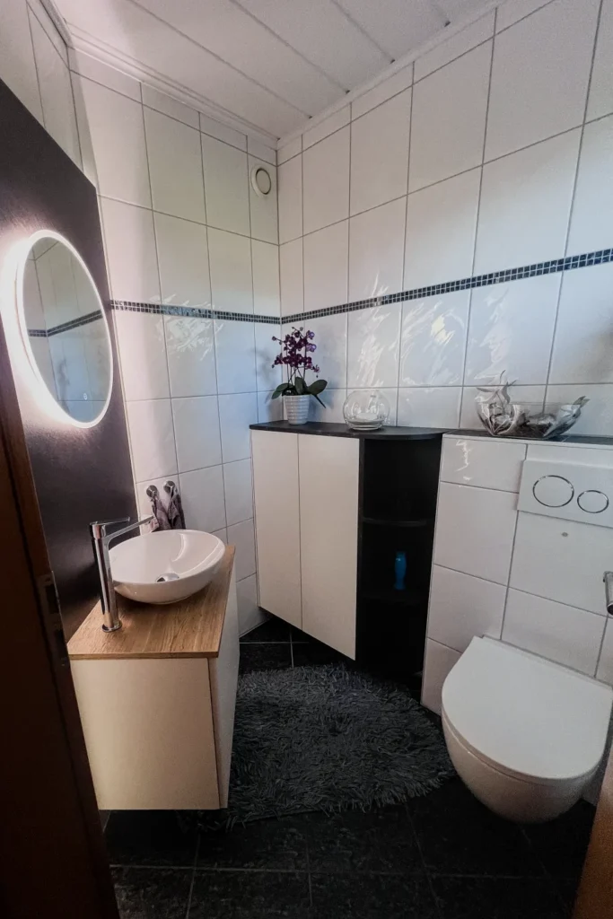 Bad mit Waschtisch, rundem Spiegel, WC und weißen Fliesen; Schrank und Ablage mit Deko.