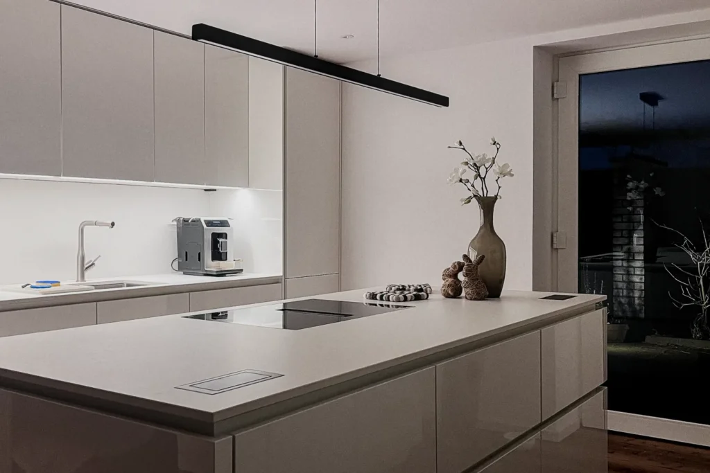 Moderne Kücheninsel mit integriertem Kochfeld, Spüle, weißen Fronten und Kaffeemaschine; Deckenlüfter über dem Kochbereich.