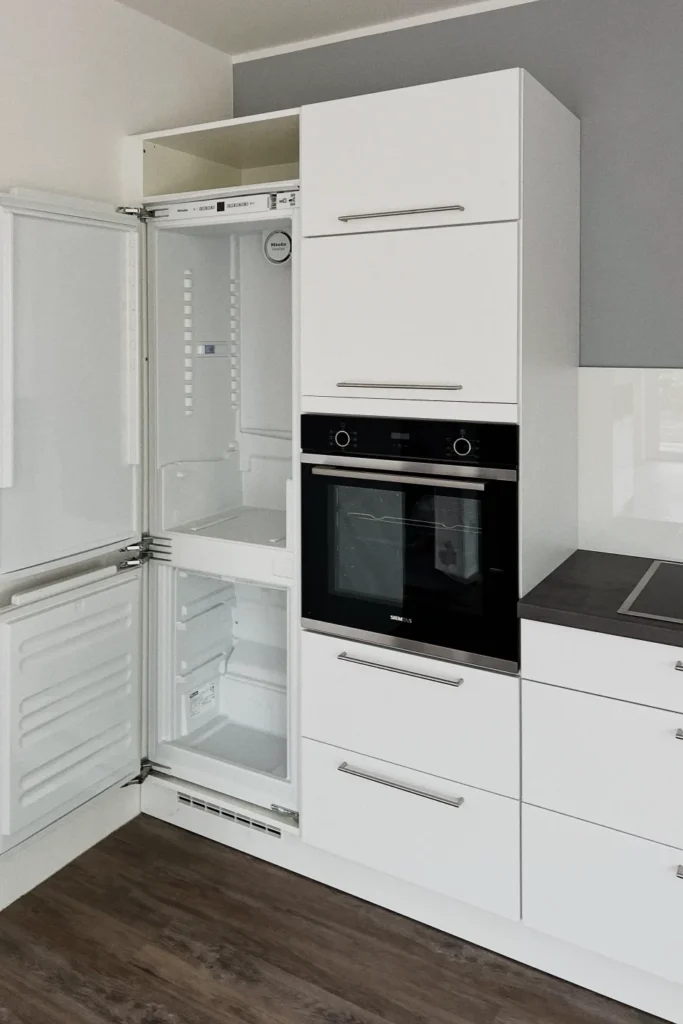 Weiße Küchenzeile mit geöffnetem Kühlschrankfach und eingebautem Backofen, sichtbar sind Fronten und Schubladen.