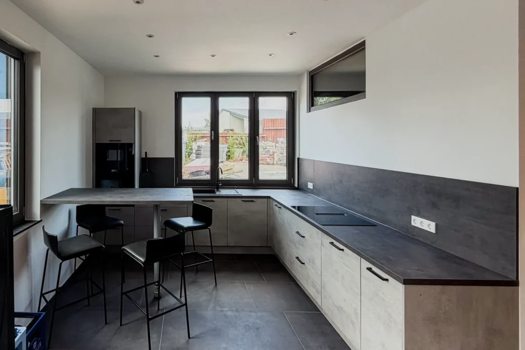 Moderne L-Küchenzeile mit Ecksitzbereich, Backofen, Spüle und großem Fenster im hellen Küchenraum.