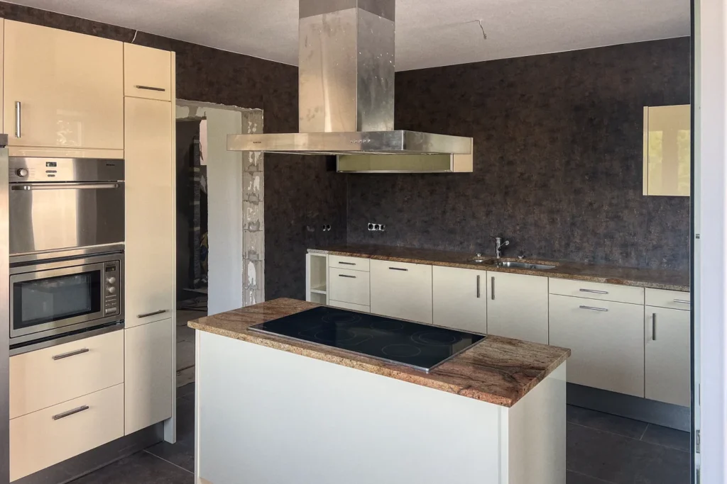 Moderne Einbauküche mit Kochfeld, Backofen, weißen Schränken und dunkler Arbeitsplatte unter Edelstahl-Dunstabzug.