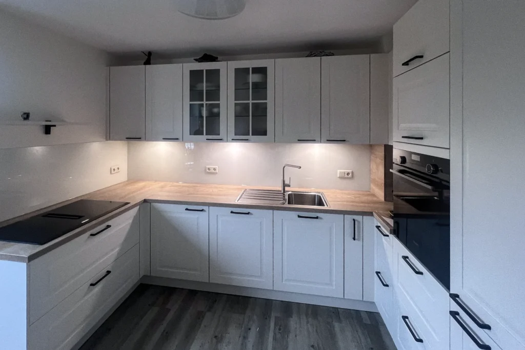 Moderne L-förmige Einbauküche mit hellen Fronten, Spüle, Kochfeld und Backofen sowie Hängeschränken mit Glastüren