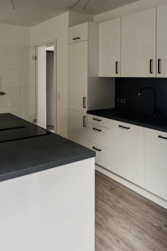 Weiße L-förmige Küchenzeile mit schwarzen Arbeitsplatten, Spülbecken und Wand-Oberschränken im modernen Design.