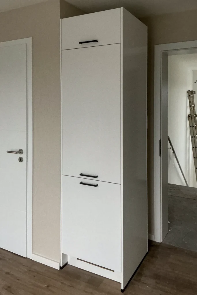 Hochschrank mit zwei Türen und Schubladen, schlichten Fronten und schwarzen Griffen, neben einem weißen Eingangstürrahmen.
