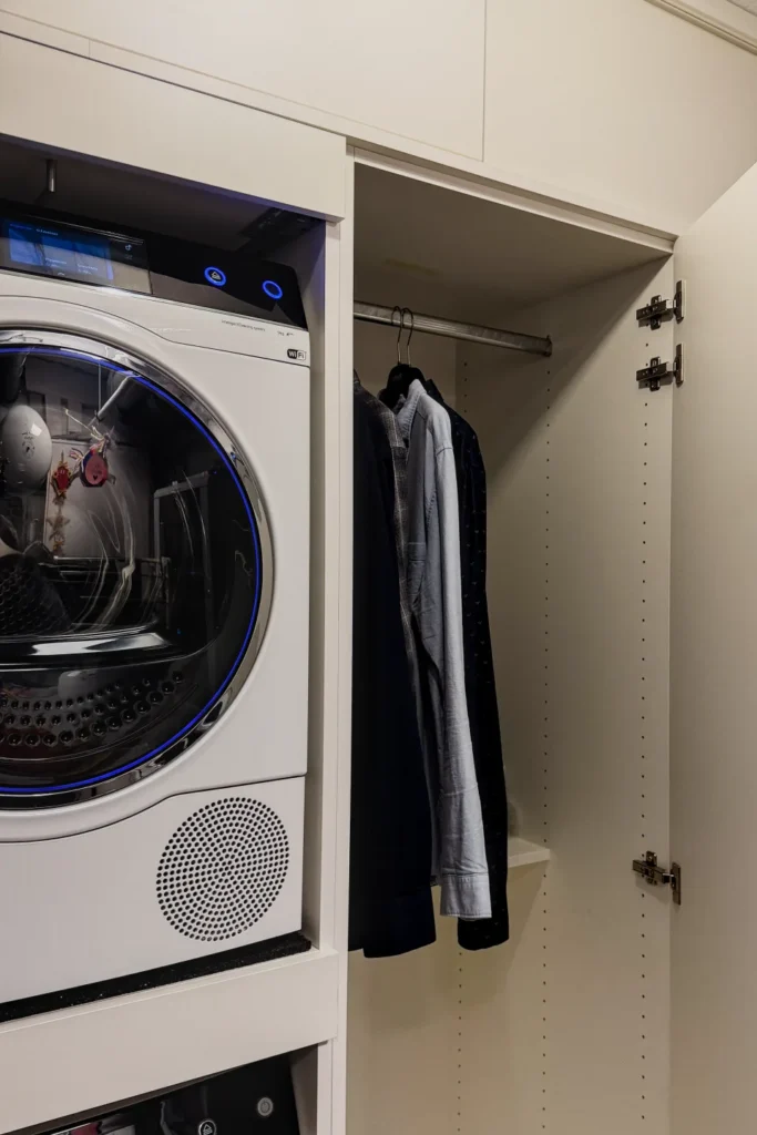 Eingebauter Wäscheschrank mit Waschmaschine und Garderobenstange, auf Bügeln hängende Mäntel.