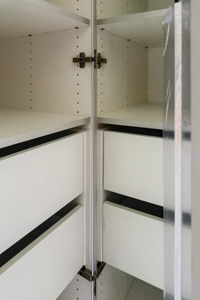 Offener Küchenschrank mit weißem Innenausbau, schmalen Schubfächern und verstellbaren Einlegeböden, Scharniere sichtbar.