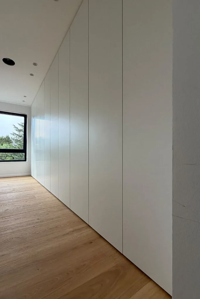Schlichtes Sideboard mit weißen Schrankfronten und senkrechten Fugen, Holzfußboden und Fenster im Hintergrund.