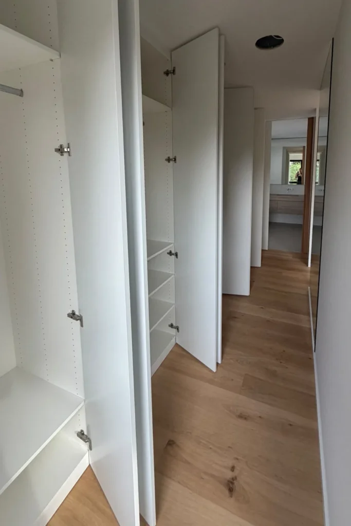 Weiße Einbauregale mit geöffneten Türen im Flur, daneben Holzfußboden und Blick in einen Wohnbereich
