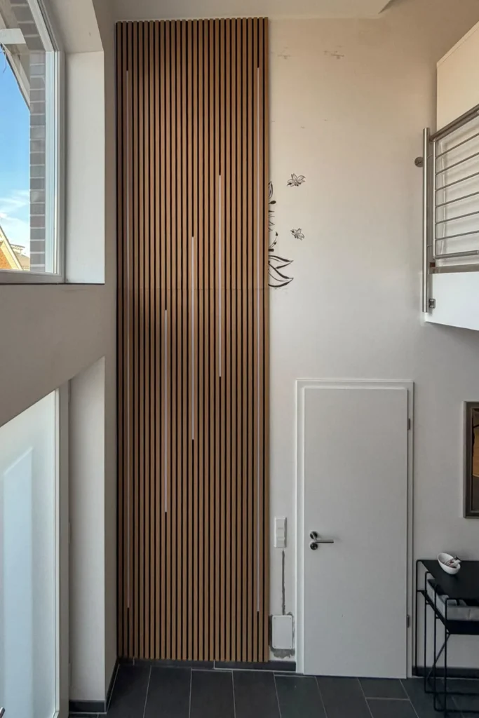Schmale Holzlamellen-Türverkleidung im Flur mit weißer Eingangstür, Fenster und schwarzem Fliesenboden